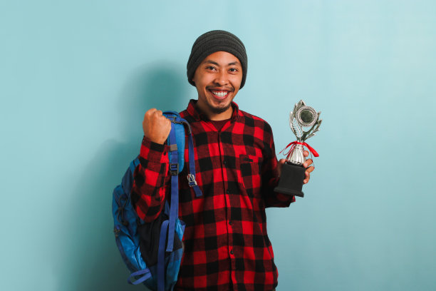 兴奋的年轻亚洲男学生拿着奖杯做出“是”的手势，孤立在蓝色背景上图片下载