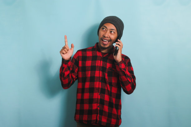 兴奋的年轻亚洲男子指着副本空间，一边讲电话，孤立的蓝色背景图片下载
