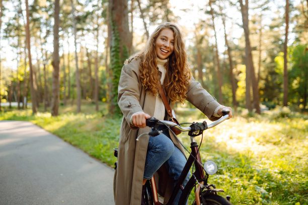 年轻女子穿着时髦的衣服，在阳光明媚的公园里骑着自行车，在户外，看向别处。图片下载
