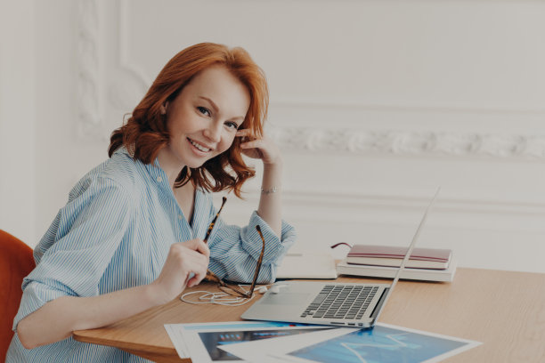 一位有着狐狸头发的满意的女商人在室内用笔记本电脑工作，在网上数据库中查看会计文件，周围是文件，在工作场所思考创意。图片下载