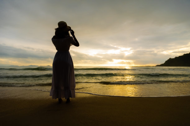 亚洲妇女的剪影戴着时尚的帽子和衣服，看着大海，享受美丽的日落在海滩上。图片下载