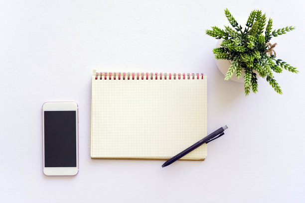 白色的桌子上放着笔记本、钢笔、智能手机和绿色植物。图片下载