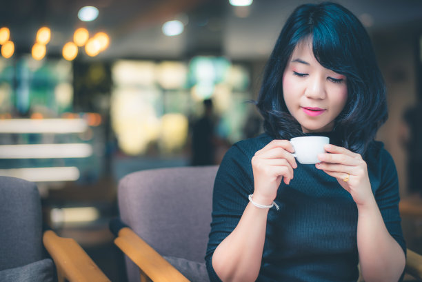美丽迷人的年轻亚洲女子手里拿着一杯咖啡，坐在咖啡馆的沙发上，早上，复古的色调。图片下载