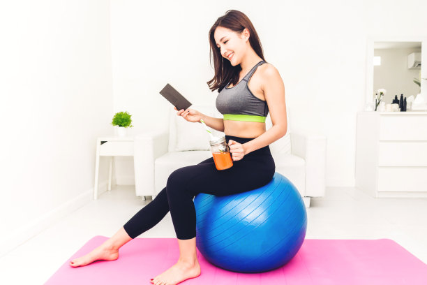 运动妇女穿着运动服坐在放松看书和喝新鲜果汁后，在蓝色健身球在家里锻炼。饮食的概念。健身和健康的生活方式图片下载