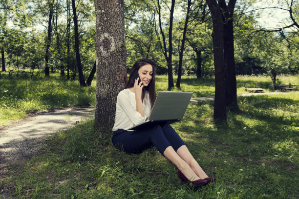 年轻的女商人坐在地上，用笔记本电脑工作，在公园里打电话选择性聚焦图片下载