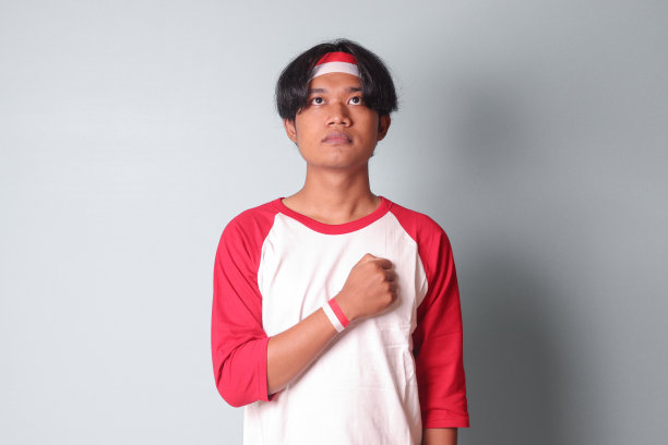 照片上的亚洲帅哥身穿t恤，头上系着红白丝带，双手放在胸前，表示敬意和敬礼。灰色背景上的孤立图像图片下载