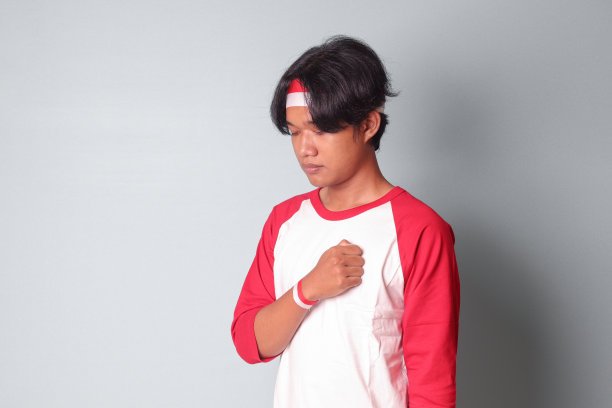 照片上的亚洲帅哥身穿t恤，头上系着红白丝带，双手放在胸前，表示敬意和敬礼。灰色背景上的孤立图像图片下载