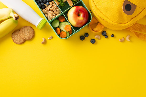 营养与创造力在这个学校餐概念中相遇:俯视图是一个午餐盒，里面装着健康的食物、酸奶瓶和一个黄色表面上的有趣背包图片下载