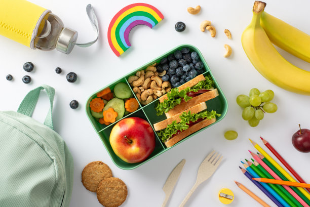 这是一张令人愉快又注重健康的午餐照片，午餐盒里有开胃的三明治，充满活力的水果和蔬菜，白色孤立的表面上有文具和背包，文字或广告的文案空间图片下载