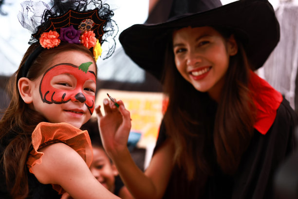 万圣节派对。一个小女孩穿着野猫服装，脸上化着妆，在万圣节派对上玩得很开心。给孩子们画脸。年轻女子在女孩脸上涂上颜料，为万圣节派对做准备图片下载