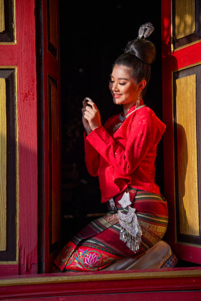 垂直形象可爱的亚洲泰国妇女穿着泰国兰纳传统风格，坐在古建筑的红色门口，幸福地看着手镯或装饰品。图片下载