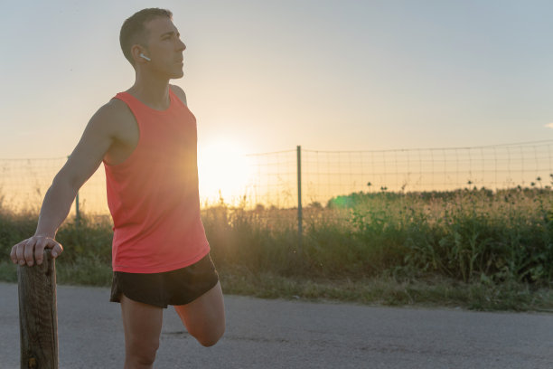 年轻的跑步者做伸展腿，他的手倚在一根木杆上，夕阳在他的身后。图片下载