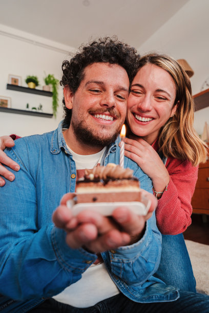 幸福的年轻夫妇庆祝生日的垂直肖像，小蛋糕吹蜡烛。妻子在结婚周年纪念日给丈夫一个有趣的惊喜，同时从背后拥抱他图片下载