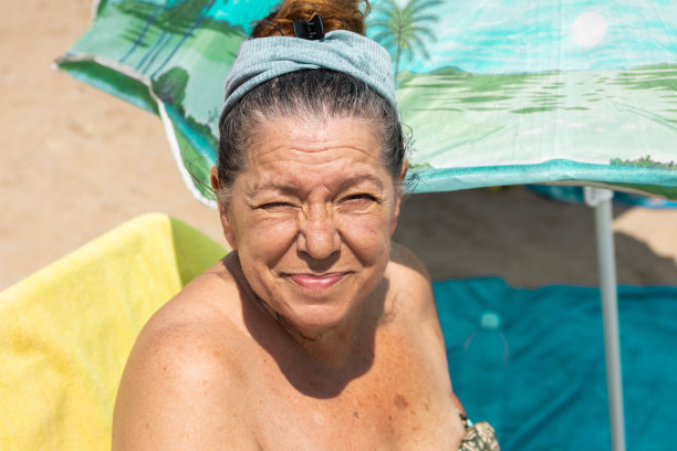 一位年长的成年妇女，在阳光下闭着眼睛，坐在吊床上，撑着伞，躺在沙滩上。概念的阳光和暑假图片下载