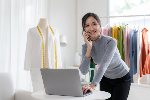服装老板在打电话。美丽的亚洲在线企业主，他们在网上销售布料，并使用笔记本电脑解释产品的特点，价格和特点。图片下载