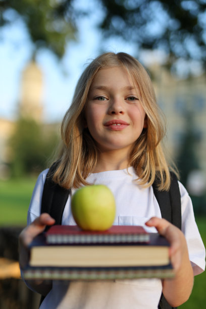 一个小学生背着书包，拿着苹果，在有钟楼的教学楼前拿着书摆姿势。图片下载