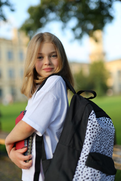 一个快乐的女学生背着圆点背包和一本红色的书在公园里享受夏日。图片下载