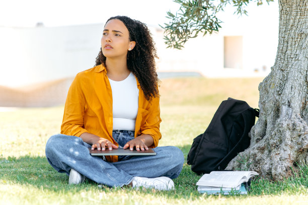 一个沮丧失望的巴西或西班牙卷发女孩的全身照片，自由职业者，坐在树旁的草地上，带着一台关闭的笔记本电脑，感到焦虑，沮丧，悲伤地看向别处图片下载