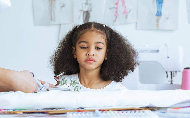 可爱的非洲小女孩拿着剪刀裁剪衣服，在裁缝室纸上设计，舒适的家是她的职业梦想，微笑着幸福和乐趣。休闲、生活、教育、儿童理念。图片下载