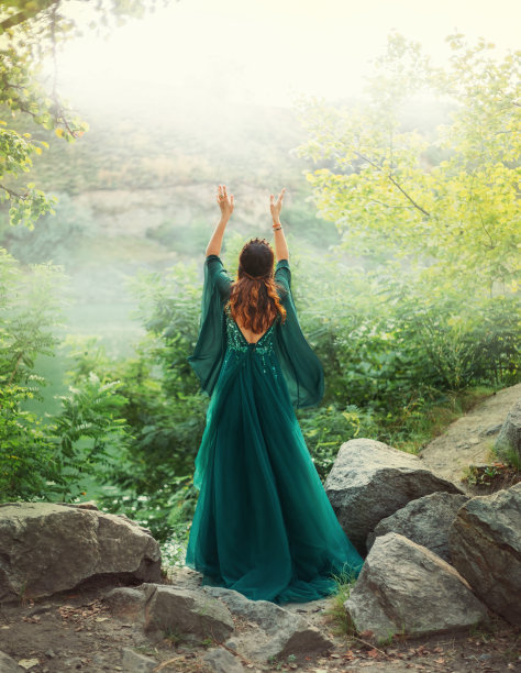 幻想中的红发女女王在夏日森林里向天空举手祈祷。女孩穿着优雅的皇家复古精灵绿色长裙。宽长袖绸在风中飞舞。艺术照片裸露开背。图片下载