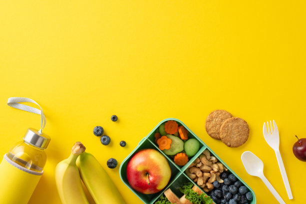 营养学校午餐盒设置。环保容器的俯视图，黄色的表面上摆放着健康的零食，坚果，浆果，饼干，水瓶，餐具，空白区域为文字图片下载