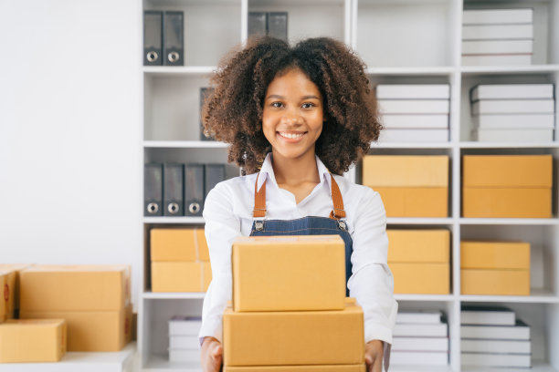 一位非裔美国女性，拿着包裹，看着镜头，站在现代办公室的几个盒子中间。图片下载