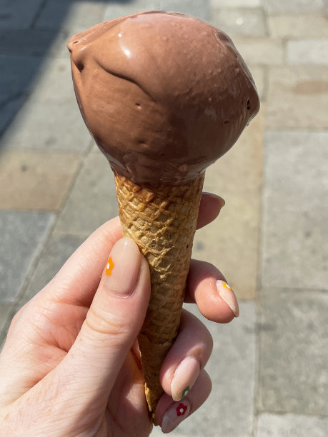 特写照片:一个无法辨认的人拿着华夫蛋筒，里面装着一勺巧克力冰淇淋，正在融化的冰淇淋，在阳光明媚的日子里走在城市街道上，聚焦在前景上图片下载