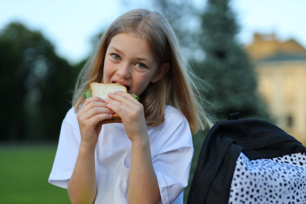 一个拿着三明治的女学生在公园里。她是一个快乐的学生，和她的背包和树一起享受午休时间。图片下载