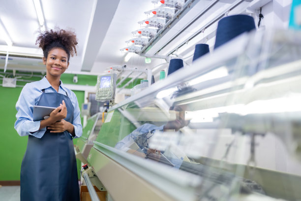 美国女设计师手持服装设计平板，面带微笑。靠近几台织布机。上面有一卷线。在一个大型的缝纫行业图片下载