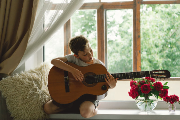 可爱的男孩在窗边的窗台上学弹古典吉他。图片下载