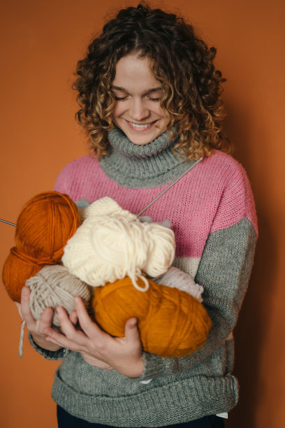 年轻的卷发女子穿着毛衣，手里拿着毛线球和针织针，在橙色的背景下摆姿势。编织，钩针，爱好。图片下载