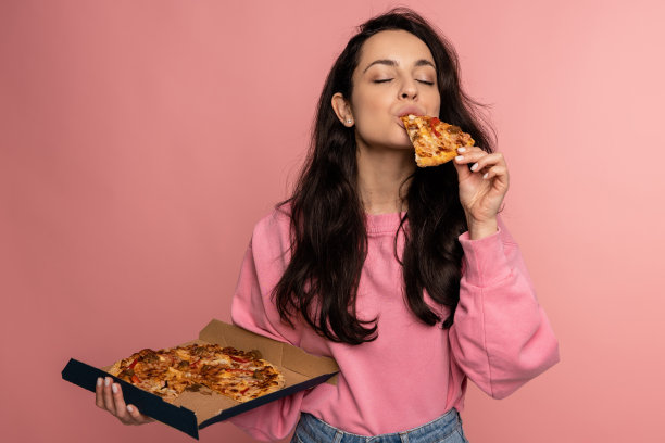 在摄影棚里拍摄的照片中，饥饿的黑发年轻女性在粉红色的背景上吃着开胃的意大利食物。不健康营养概念图片下载