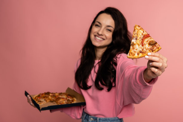 在工作室拍摄的照片中，微笑的年轻女子在粉红色的背景上摆着她最喜欢的意大利食物。不健康饮食观念图片下载