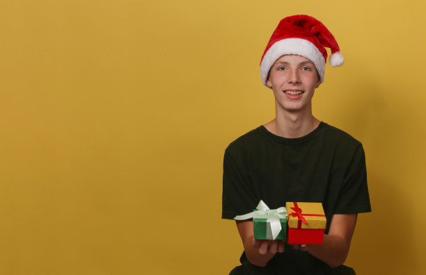 黄色背景的少年头戴圣诞老人帽，手里拿着圣诞礼物。图片下载