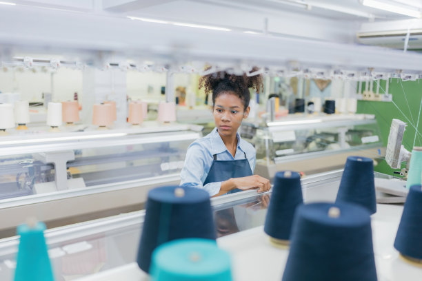 美国女设计师用你的手打开盖子，检查电脑缝纫机。在一个庞大的织造工业中，周围有线和制衣设备。图片下载
