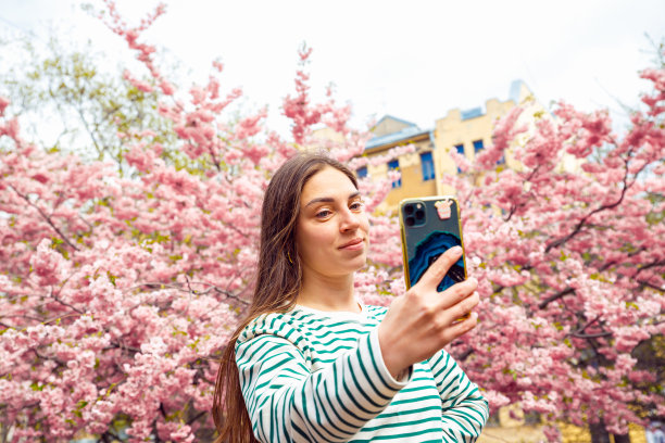 年轻女性正在用手机说话。白人女模特在春天的樱花公园用智能手机自拍。图片下载