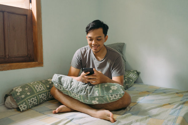 肖像亚洲男子微笑着看着智能手机，在卧室里工作，自然光的概念图片下载