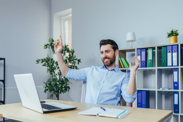 一个商人在明亮的办公室里工作，在桌子旁用笔记本电脑跳舞庆祝成功图片下载