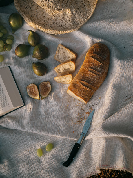 直接从上方拍摄浪漫的地中海野餐。草帽，意大利面包，无花果，绿葡萄，打开的书，撒丁岛刀，都放在一张白色的天然桌布上。图片下载