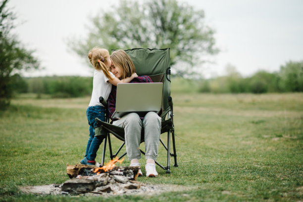 妈妈和孩子在户外上网。隔离，在冠状病毒爆发期间关闭了幼儿园。在火灾附近的笔记本电脑上与家人在线交流。在家上学，自由职业。图片下载