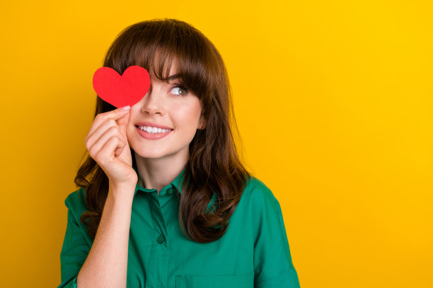 照片中梦幻般的漂亮女士穿着绿色的衬衫，心卡盖着眼睛，看着空白的空间孤立的黄色背景图片下载