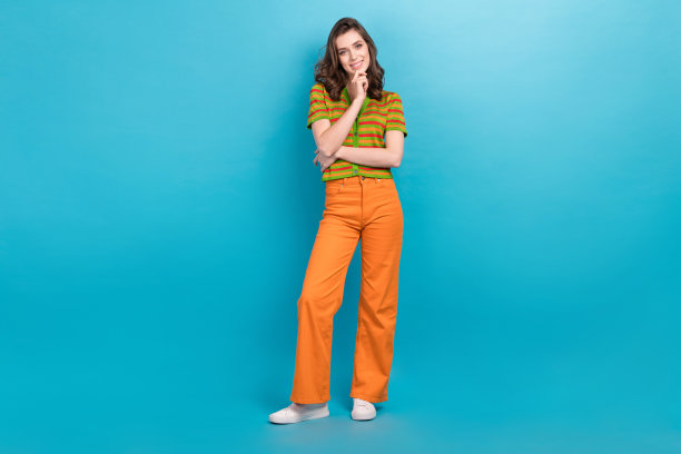 全尺寸照片，沉思的女人穿着橙色裤子，彩色开襟羊毛衫，手放在下巴上，孤立在蓝色背景上图片下载