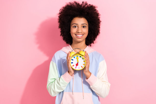 照片的漂亮快乐的小孩穿着连帽衫微笑拿着时钟孤立的粉红色背景图片下载