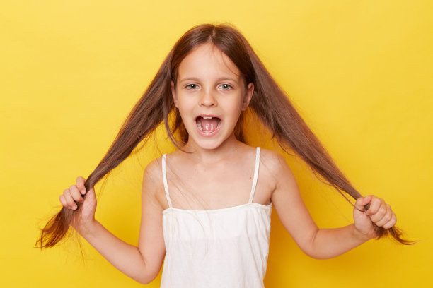 快乐兴奋的小女孩，扎着马尾辫，孤立地站在黄色的背景上，表达着兴奋积极的情绪，拉着她的头发图片下载