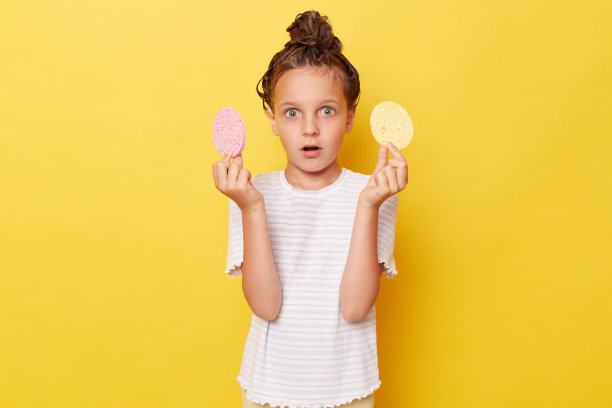小女孩头发湿漉漉的，穿着休闲的白色t恤，站在黄色的背景下，看着镜头，大眼睛拿着化妆海绵。图片下载