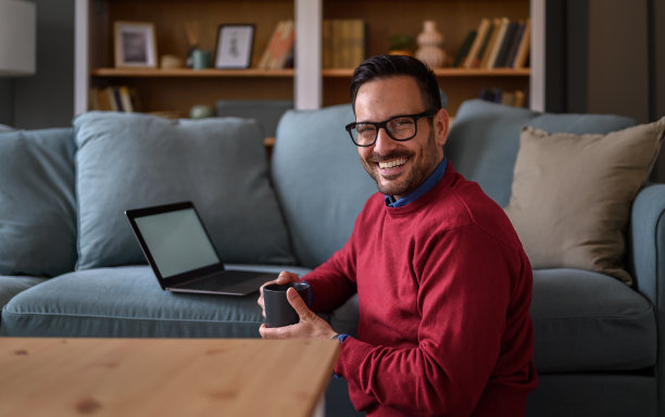 客厅里，年轻的商人拿着咖啡杯坐在笔记本电脑和沙发旁，面带微笑图片下载