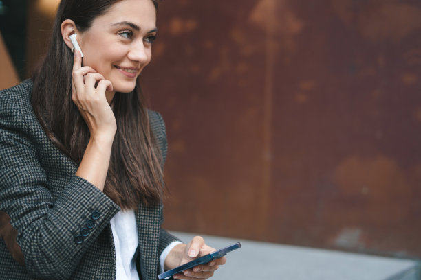 漂亮的黑发年轻女子穿着休闲夹克使用电话和无线耳机。人们远程工作概念图片下载
