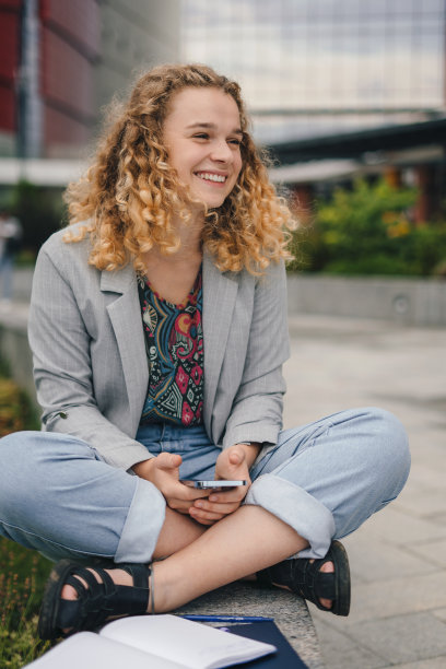 一位穿着夹克、面带微笑的女士在校门外用手机望向别处。技术的概念。图片下载