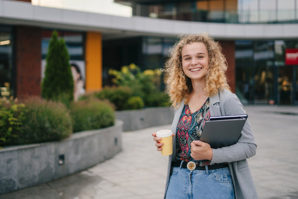 在城市里，卷头发的女人拿着外带的咖啡因饮料和关闭的笔记本电脑。聊天，一起工作，微笑，时髦的自由职业者，拿着笔记本电脑图片下载