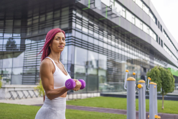 漂亮的粉色头发的健身女人在公司大楼附近的公园里举哑铃。图片下载
